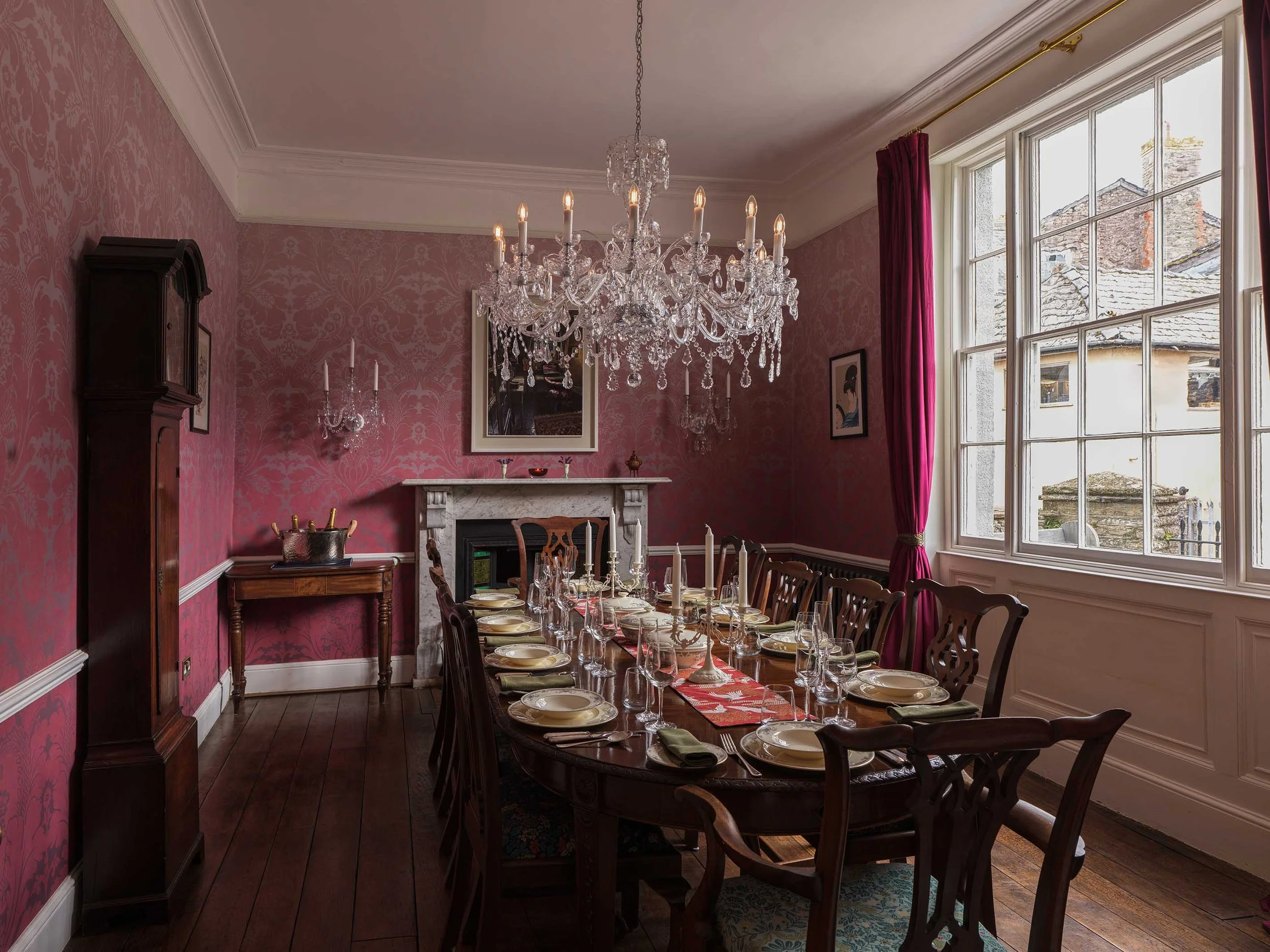 Elegant dining room with red wallpaper, large window with red curtains, wooden table set for a formal dinner with plates, glasses, and silverware, Crystal chandelier hanging from ceiling, fireplace with a mirror above, and antique furniture.