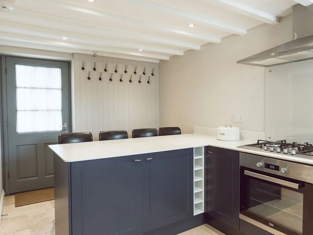 Modern kitchen with dark blue cabinets, white countertop, stainless steel oven and stove, white walls, and a door with frosted glass window. There are five black chairs along the countertop, a white cake tin nearby, and hooks on the wall.