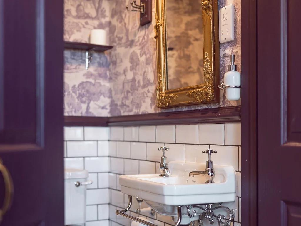 A vintage-style bathroom with a white porcelain sink, chrome fixtures, white subway tiles, a gold ornate framed mirror, and purple walls with wallpaper pattern.