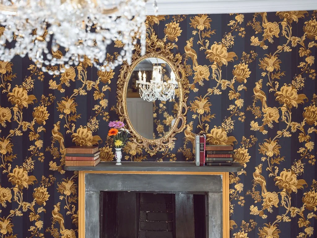 An ornate room with gold and black floral patterned wallpaper, a mirror with a gold frame above a fireplace, a chandelier reflecting in the mirror, a small vase of colorful flowers, and a stack of books on the mantle.