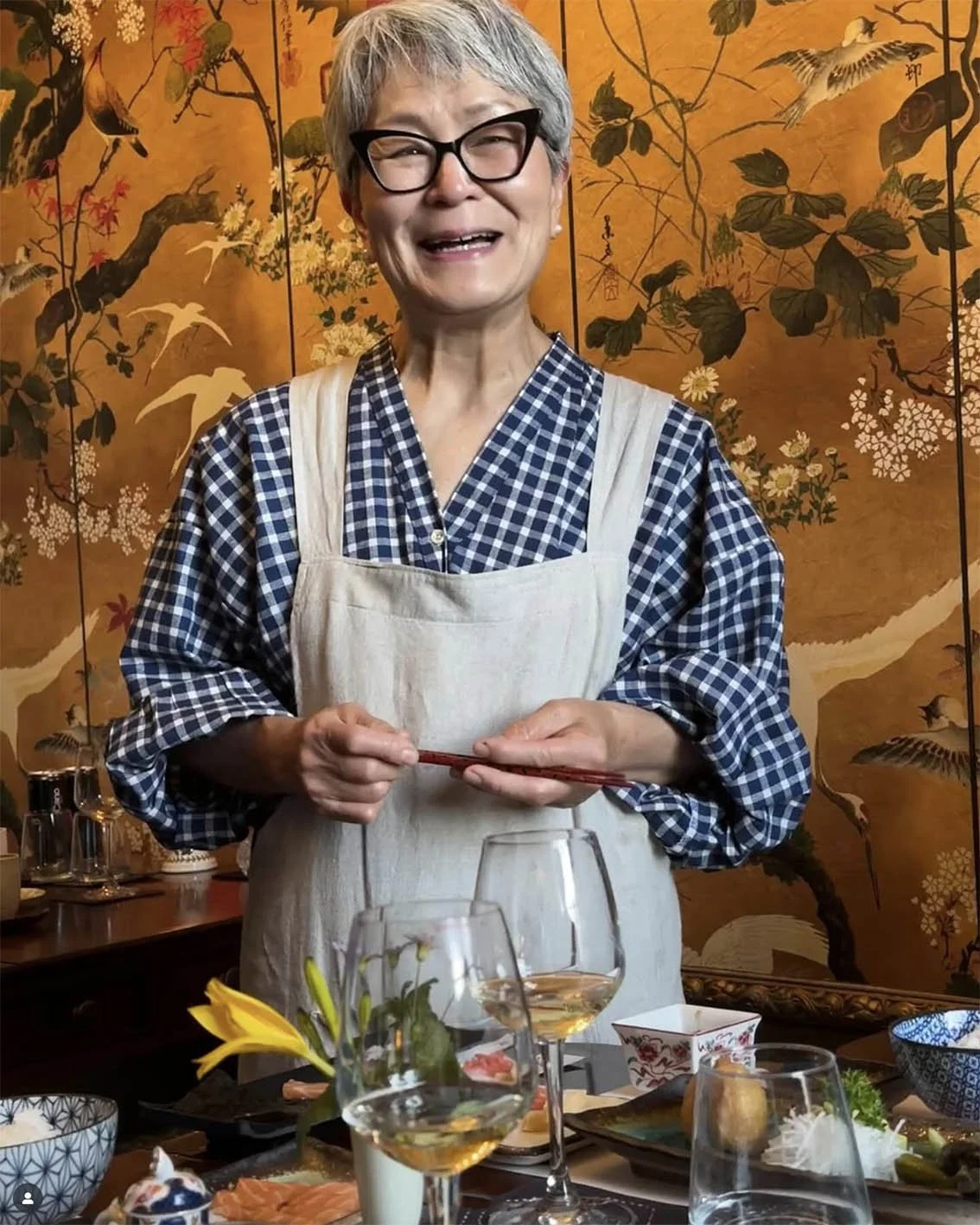 A woman with short gray hair, wearing glasses, a blue checkered shirt, and a beige apron, standing behind a table with food and drinks, smiling at the camera, with a decorative Asian-style folding screen with floral and bird motifs in the background.