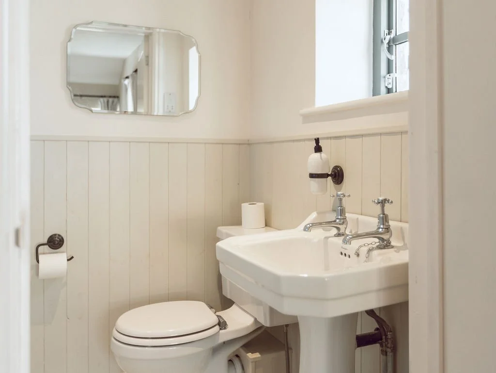 Small bathroom with a white toilet, a vintage white sink with separate hot and cold taps, a wall-mounted soap dispenser, a roll of toilet paper, a mirror above the sink, and a window.