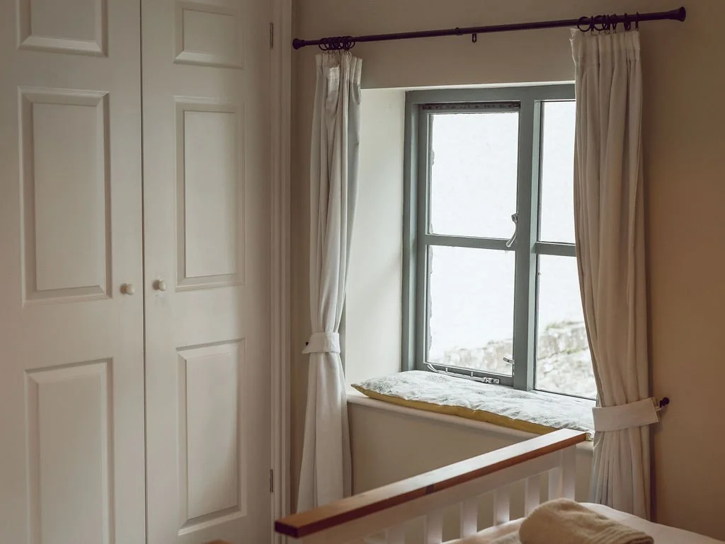 A bedroom corner with a white wardrobe, a window with white curtains, and a window seat with a yellow cushion.