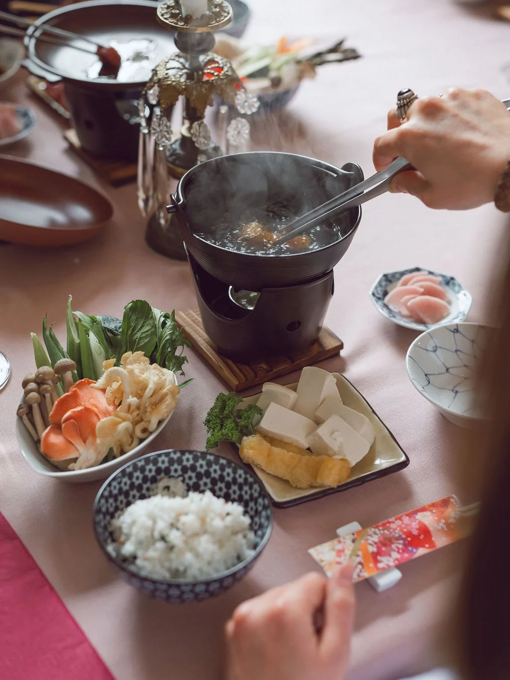 A traditional Japanese hot pot meal on a table, with various vegetables, tofu, rice, and a steaming pot in the center, with a person using tongs to add ingredients to the hot pot.