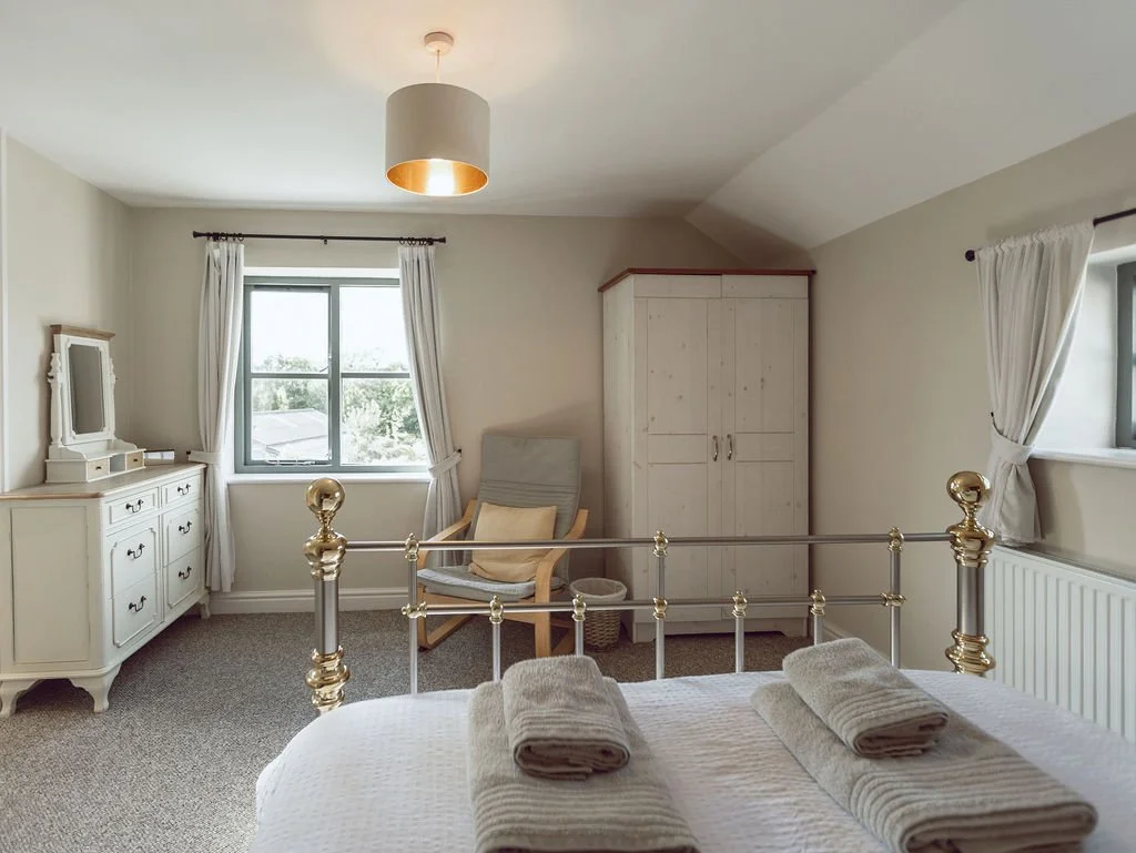 A cozy bedroom with a bed, white curtains on the windows, a white dresser with a mirror, a gray armchair with a pillow, a white wardrobe, and a ceiling light fixture.