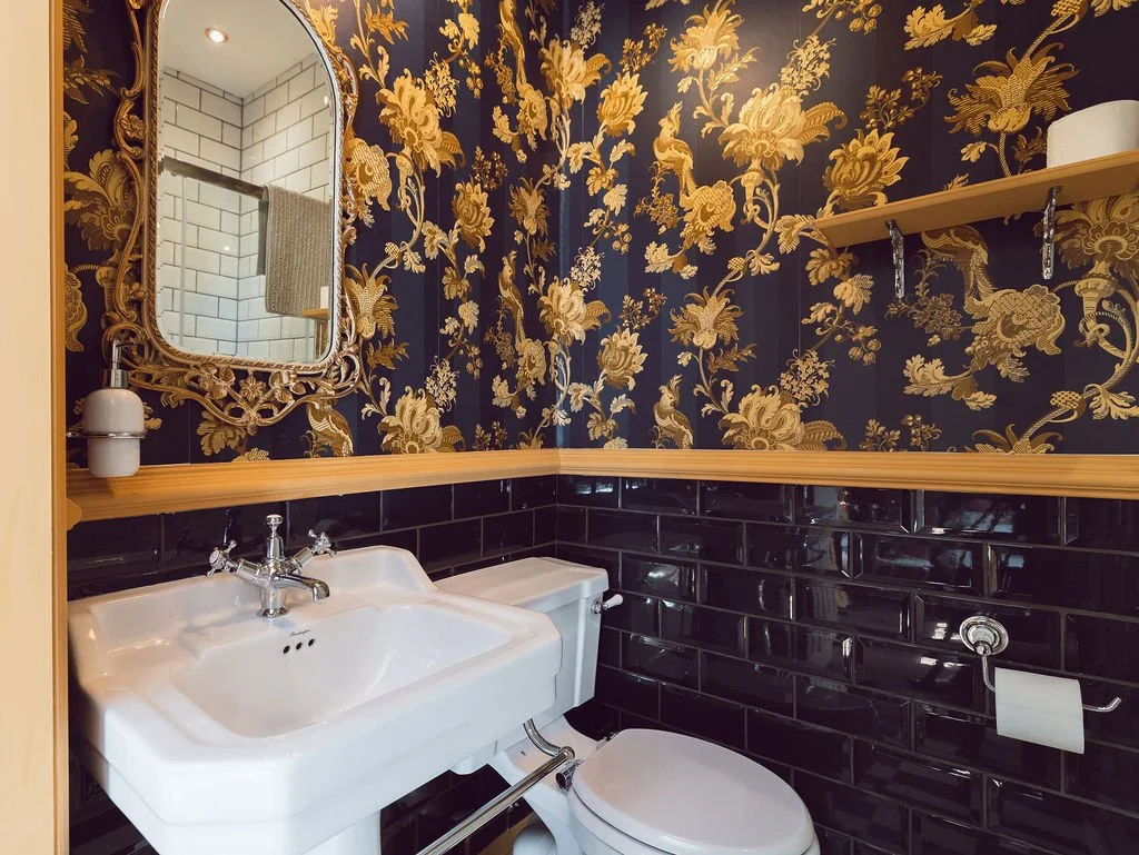Bathroom featuring a white sink with a vintage-style faucet, black subway tile walls, gold floral patterned wallpaper, a mirror with an ornate gold frame, and a wooden shelf with a white container and a roll of toilet paper.