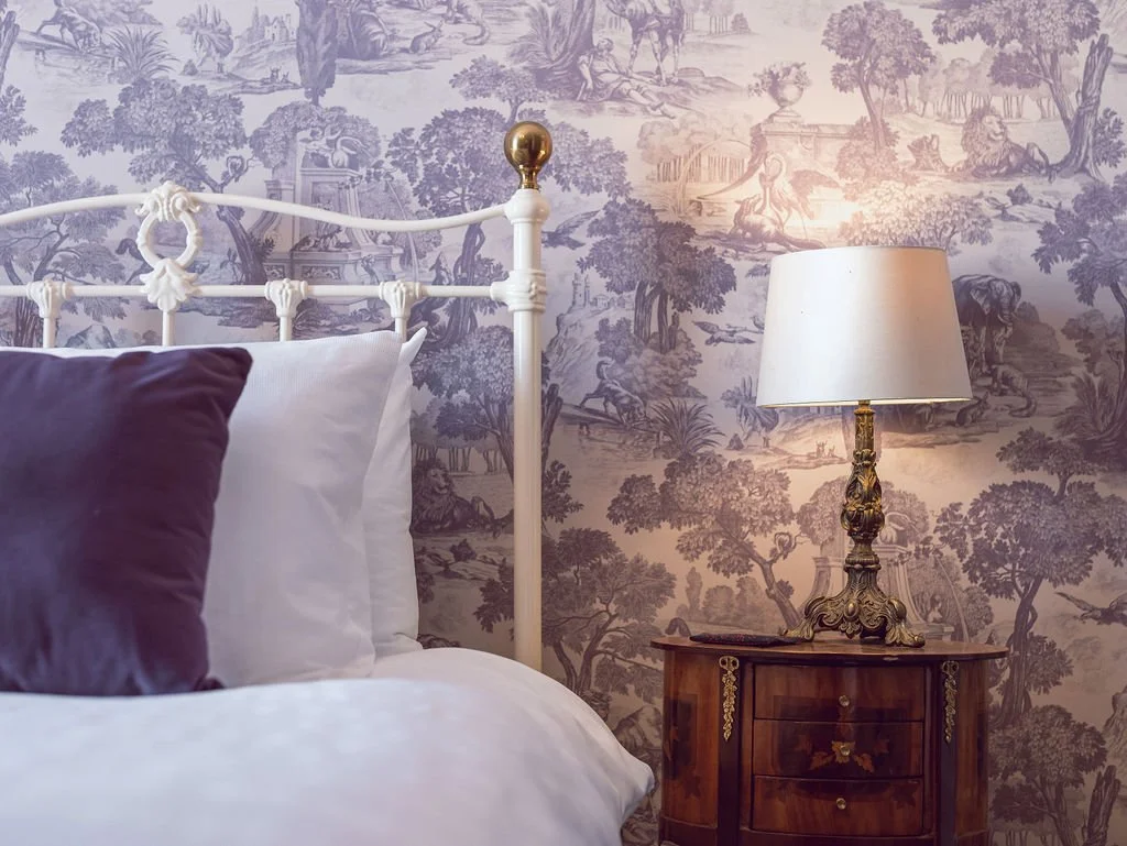 A bedroom with a white metal bed frame, purple and white pillows, a wooden nightstand, and a table lamp with a decorative bronze base, all against a wall with a scenic toile wallpaper featuring trees and landscape scenes.