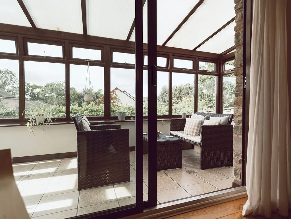 A sunroom with large glass windows and ceiling, wicker furniture including a chair and a loveseat with cushions, a small table, and potted plants, overlooking a backyard and trees.