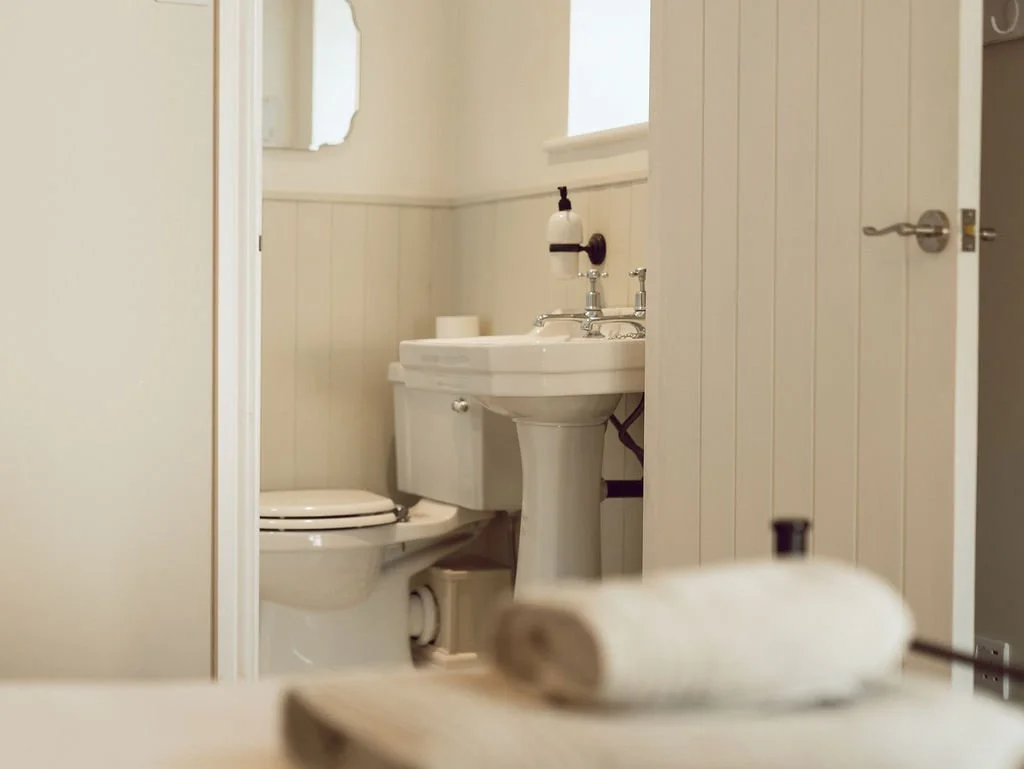 A small bathroom with a toilet and a pedestal sink seen through an open door. A mirror and a window are visible on the wall above the sink, with a roll of toilet paper on the back of the toilet and another on the sink.