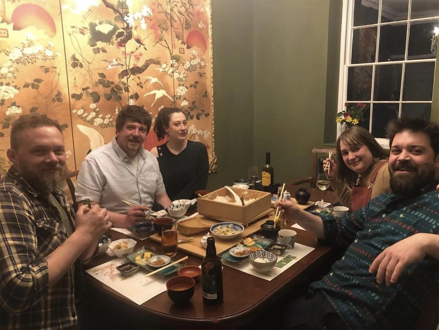 Six people sitting at a dining table with various dishes, chopsticks, wine, and beer bottles, enjoying a meal together in a cozy restaurant with Asian-themed decor and a large window.