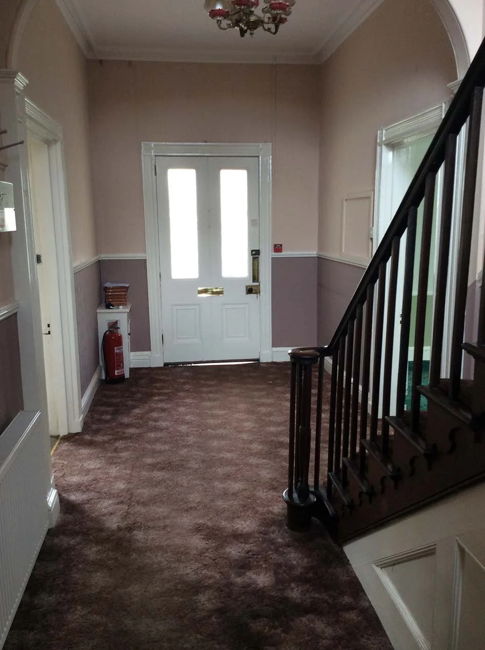 Entryway with a wooden staircase on the right, a door with frosted glass on the back wall, a chandelier on the ceiling, and a fire extinguisher on the left side.