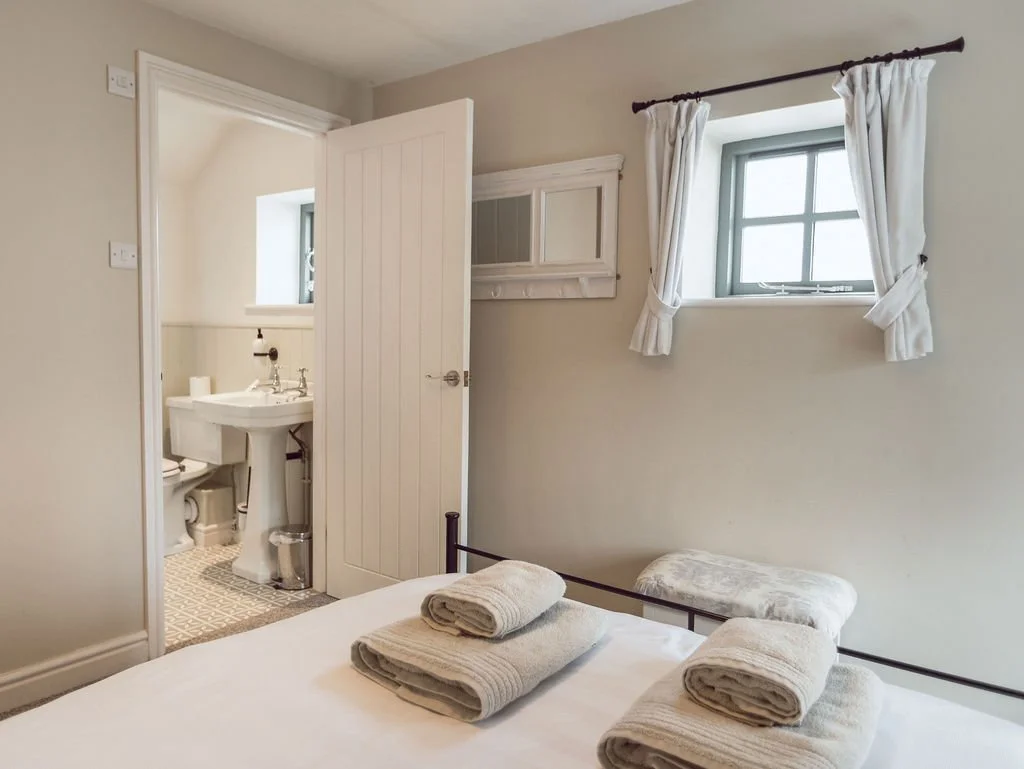 A cozy bedroom with a bed having neatly folded towels, a small bench, a window with white curtains, and an open door leading to a small bathroom with a pedestal sink and toilet.