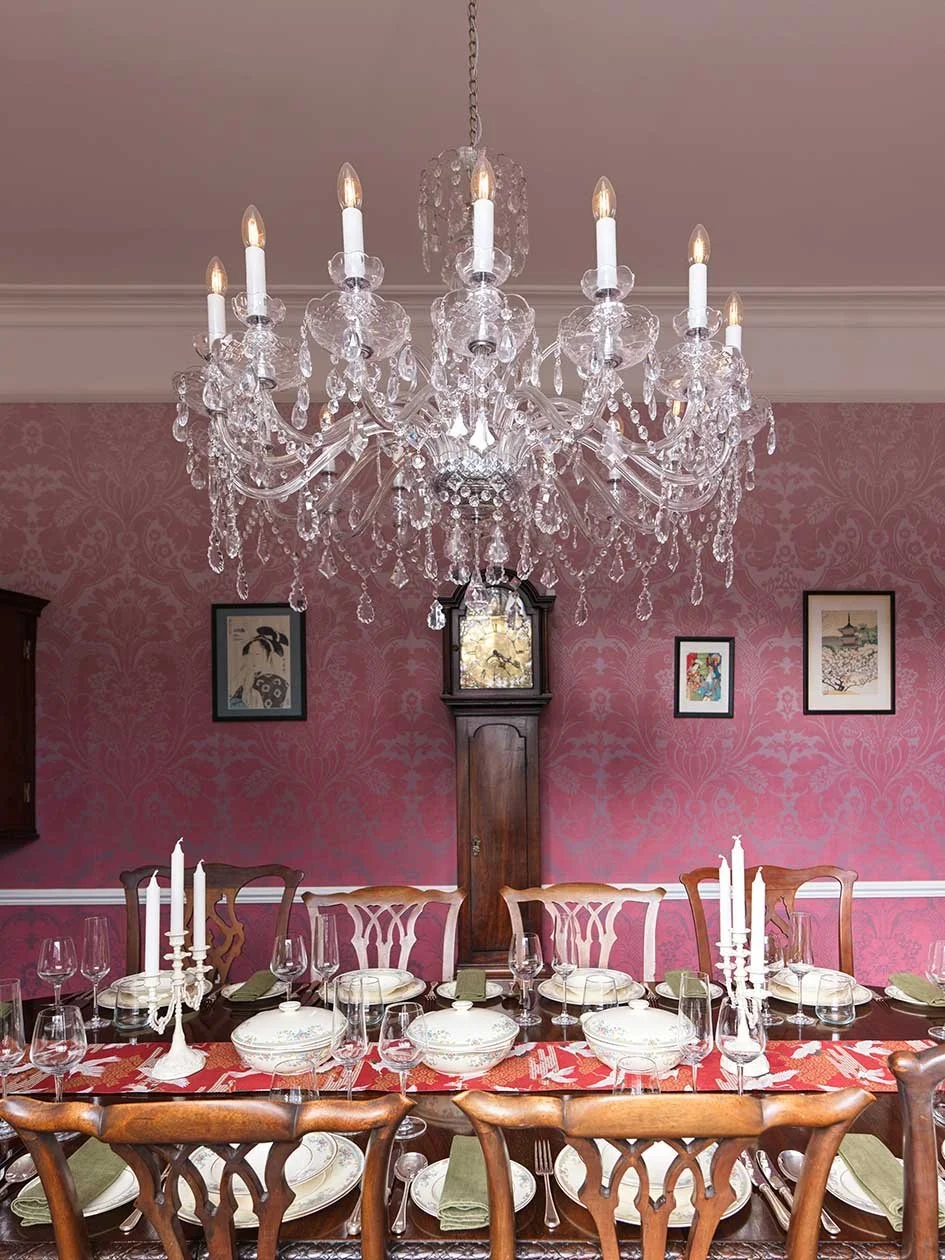 Elegant dining room with a large crystal chandelier, a long wooden table set with white and patterned dishes, glassware, candlesticks, and green napkins, against a pink damask wallpaper with framed artwork.