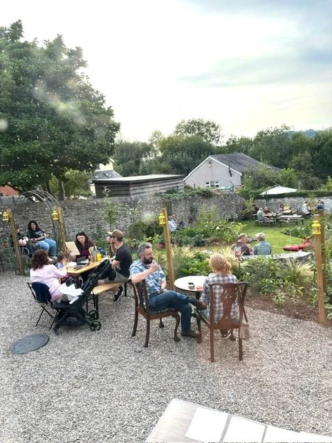 People sitting at outdoor tables and chairs in a garden, dining and socializing on a cloudy day with trees and houses in the background.