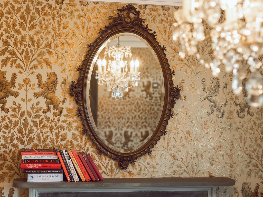 An ornate oval mirror with a dark carved frame hangs above a fireplace mantel. The mantel has a small stack of books on the left side. The wall behind features gold and beige wallpaper with a pattern of lions and floral motifs. The reflection in the 