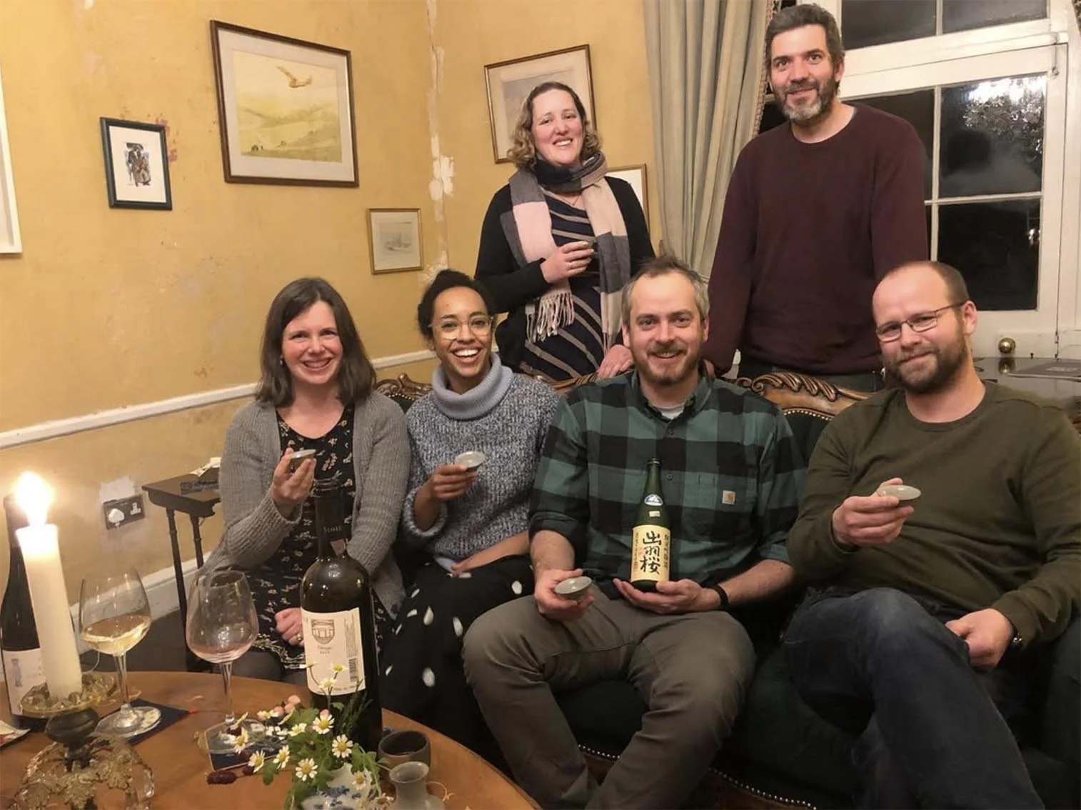 Group of six friends celebrating indoors, holding drinks, with a table of wine, a candle, and flowers in the foreground, and framed artwork on yellow walls in the background.