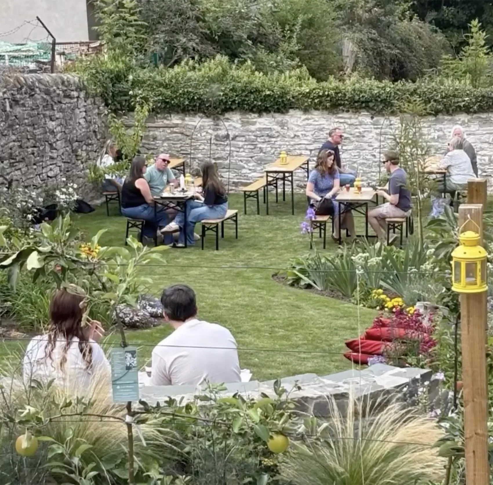 People sitting and socializing in a garden patio with tables, chairs, and lush greenery.