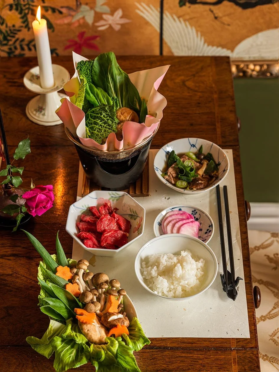A traditional Japanese meal with rice, raw meat, fresh vegetables, and pickled radish, arranged on a wooden table with a candle and floral decorations in the background.