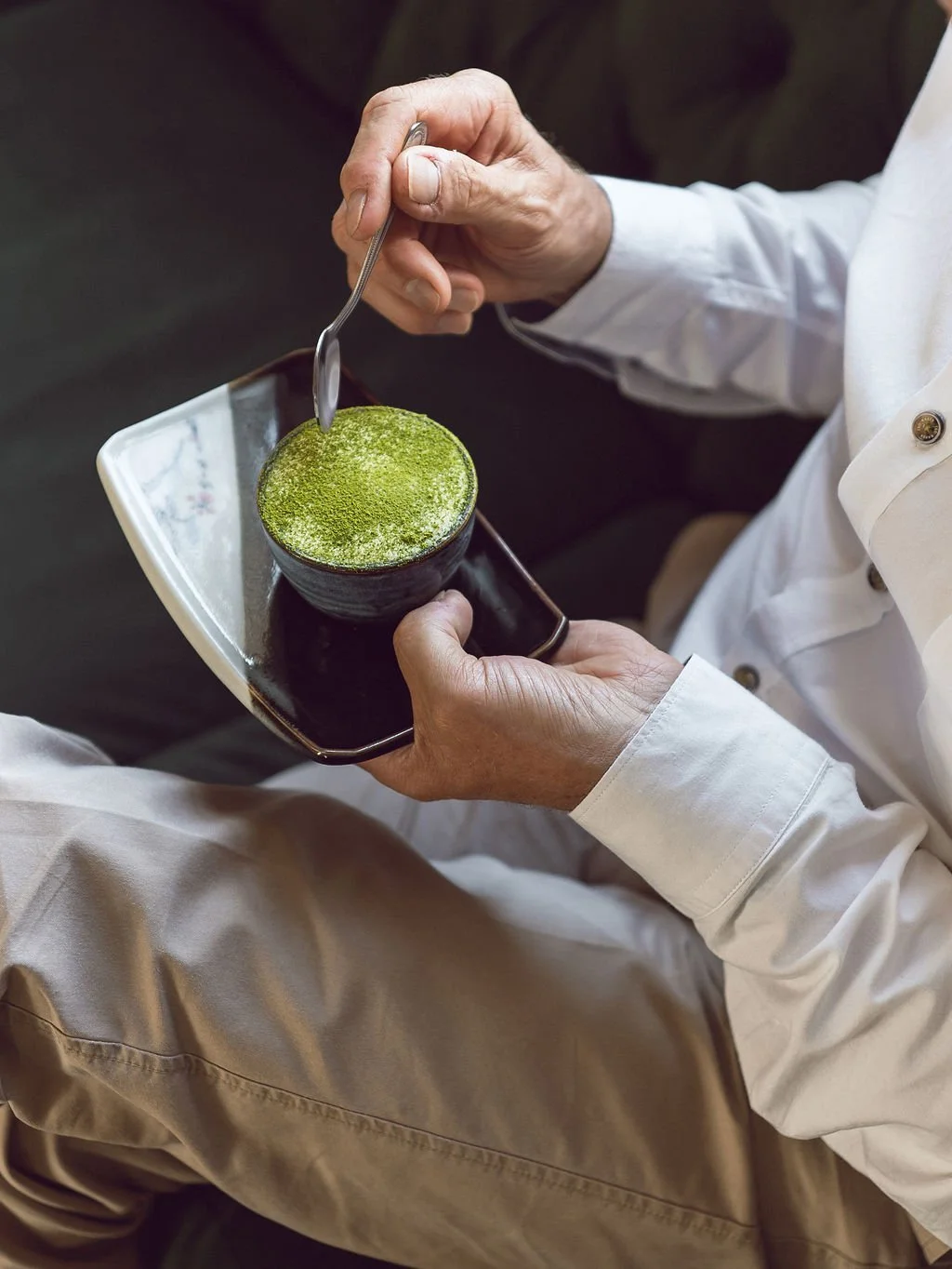 Person holding a small black bowl with matcha green tea powder and a spoon, dressed in white shirt and beige pants.