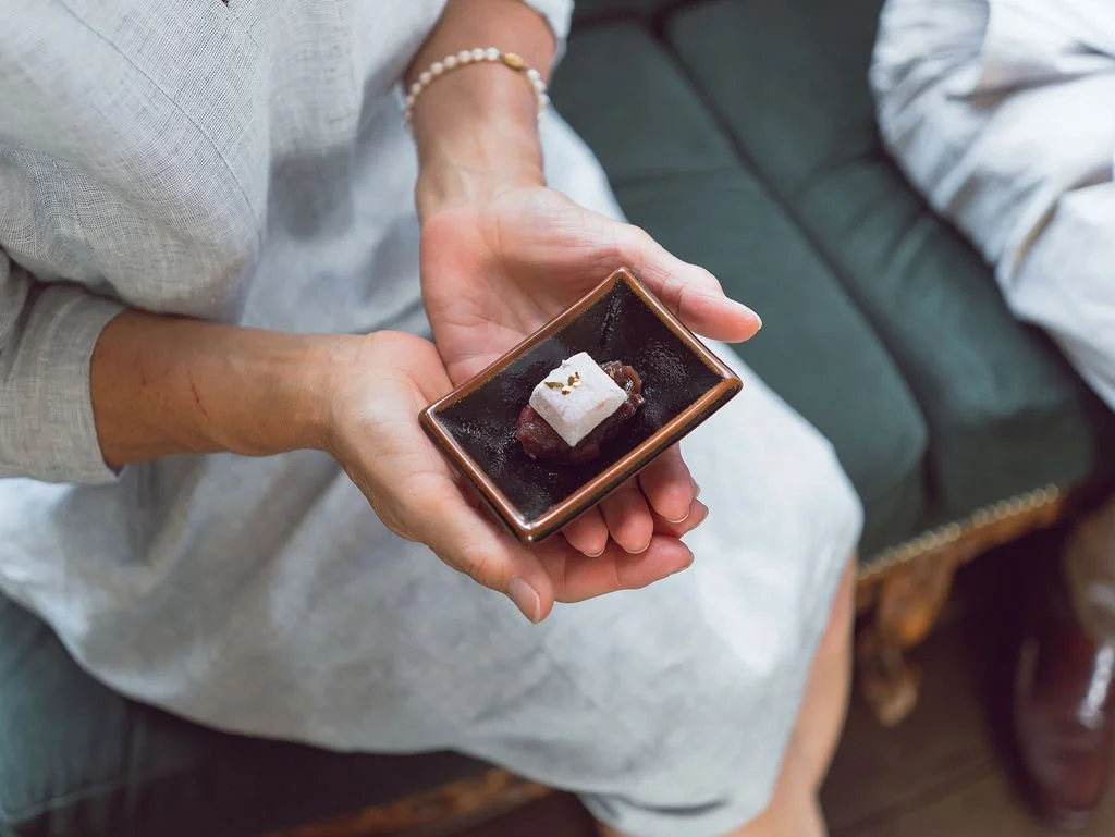 A person holding a small square dish with a piece of dessert, possibly a mochi or rice cake, topped with a small amount of gold leaf. The person wears a pearl bracelet and is sitting on a cushioned seat.