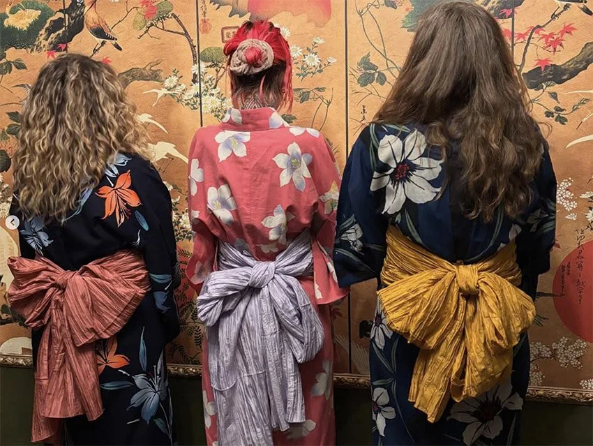 Three women viewed from behind wearing traditional Japanese kimonos with large bows on their backs, standing in front of a decorative Japanese-style screen with floral and nature motifs.