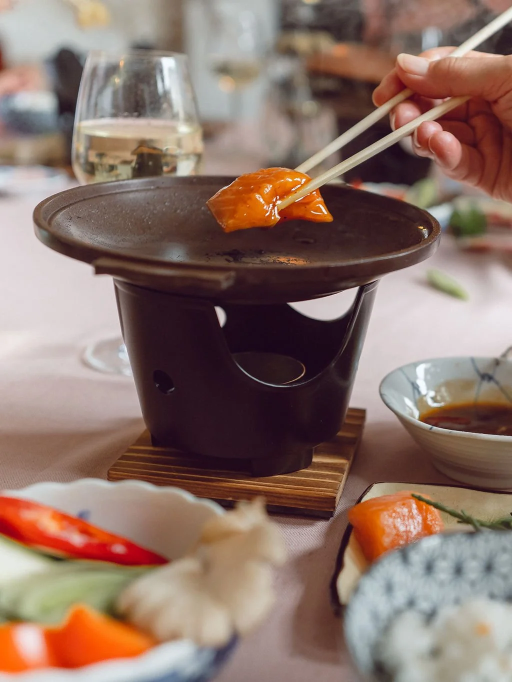 Person using chopsticks to dip a piece of sushi into a small grill or heating unit on a table in a restaurant. A glass of white wine, bowls of dipping sauce, and various dishes of sushi are visible.