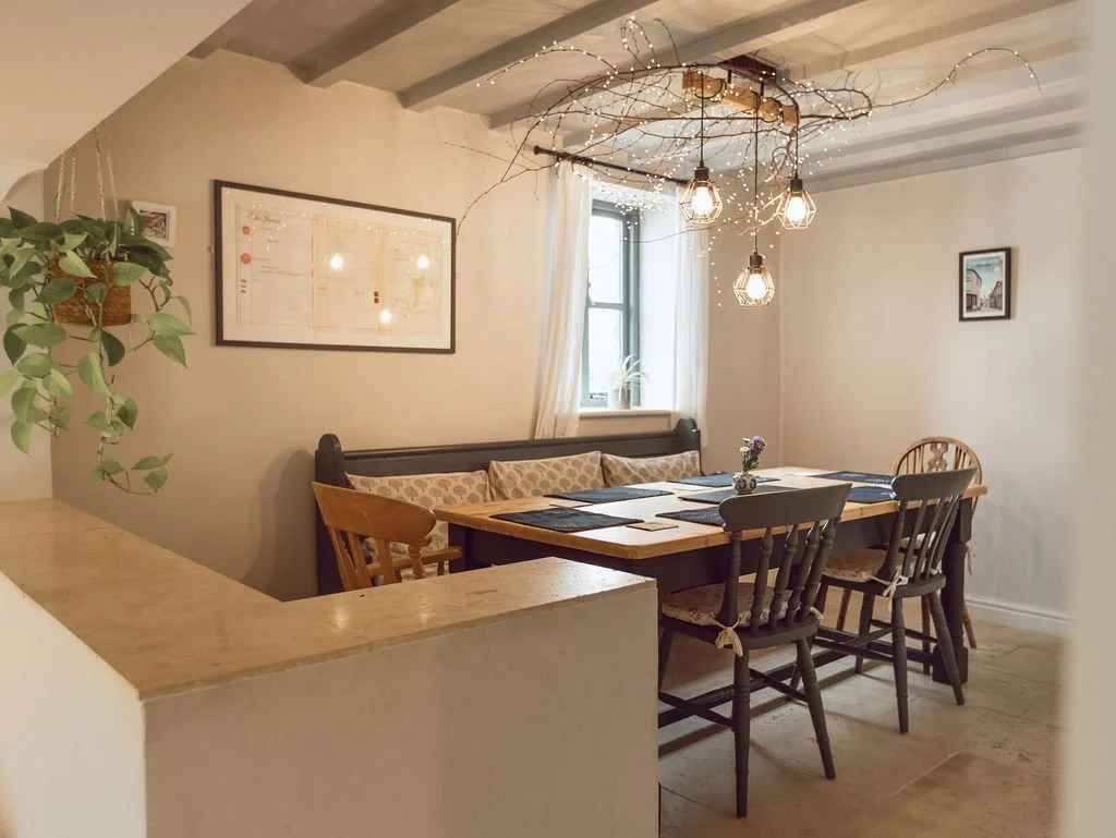 A cozy dining room with a wooden table set for six, surrounded by mixed wooden and pushed-back chairs, a bench with cushions under a window, and a decorative branch chandelier with hanging lights on a ceiling with pastel striped beams. There's a fram