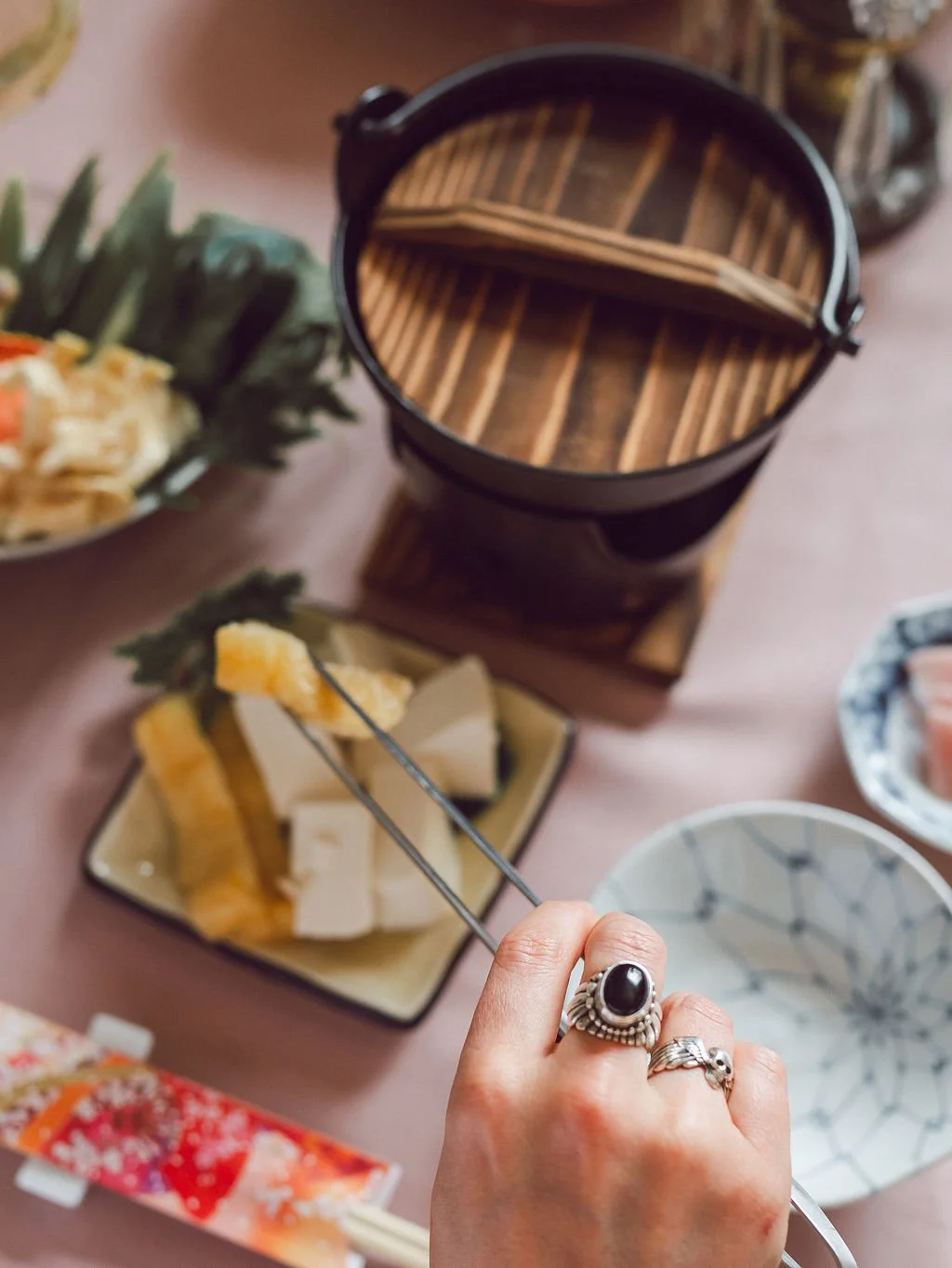 Person picking cheese with tongs at a samurai-themed dining table, surrounded by plates of food, chopsticks, and a wooden sushi boat.