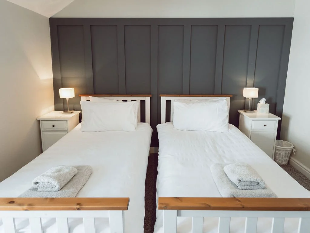 A bedroom with two twin beds with white bedding, wooden headboards, and gray towels on each bed. There are white nightstands with lamps on each side of the beds, a tissue box on the right nightstand, and a small laundry basket in the corner. The head