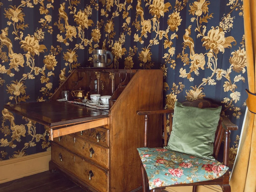 Vintage wooden tea cabinet with cups and teapot, beside an ornate floral upholstered chair with a green pillow, in a room with dark blue and gold floral wallpaper and gold curtains.