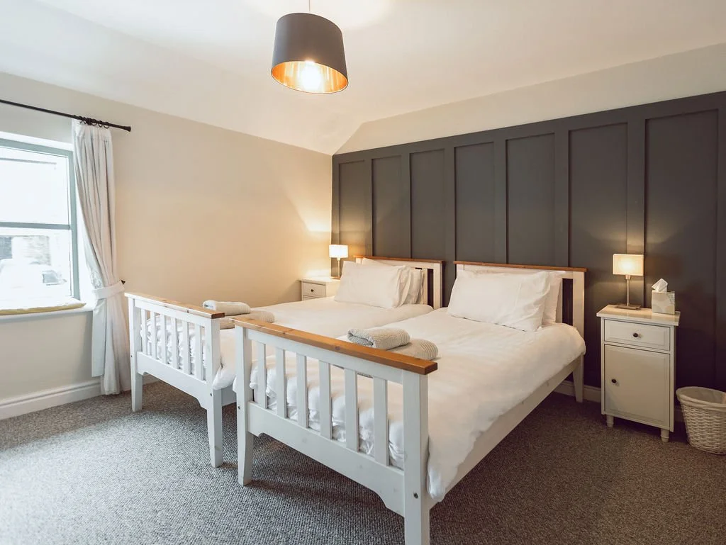 A clean, modern bedroom with two white beds, each with a dark blue headboard, nightstands with lamps, a window with white curtains, and a ceiling light fixture.