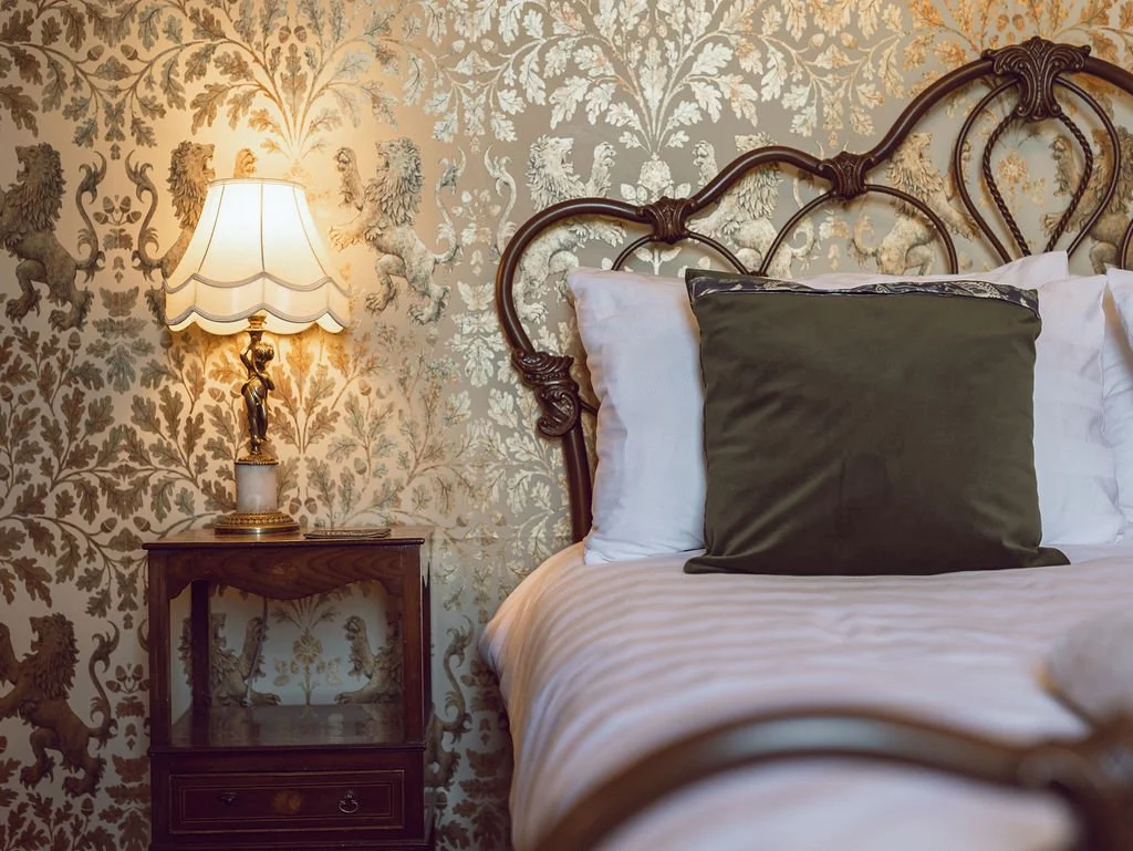 A vintage bedroom scene featuring a bed with white sheets and pillows, a dark green cushion, a decorative bronze headboard, a wooden nightstand, and a lamp with a ornate base and cream shade, set against a floral wallpaper with lion motifs.