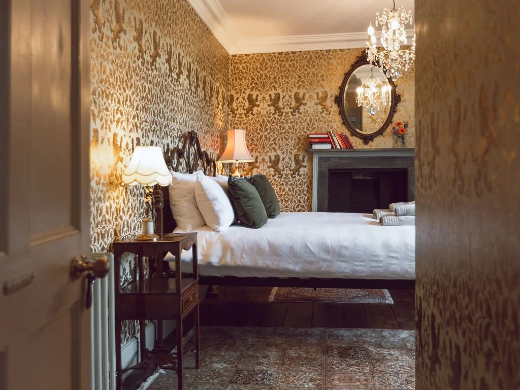 A bedroom with ornate gold wallpaper, a white bed with multiple pillows, a small side table with a lamp, a fireplace with books and a mirror above, and a chandelier hanging from the ceiling.