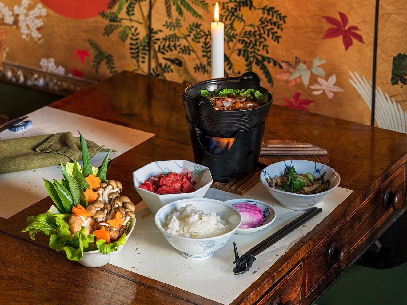 A traditional Japanese hot pot meal with various ingredients on a wooden table, including fresh vegetables, sliced meat, rice, and dipping bowls, with a small portable stove heating the hot pot.