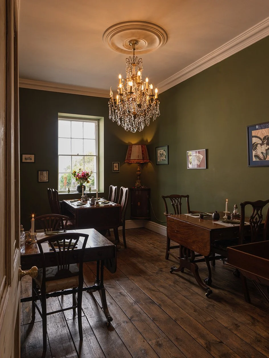 A cozy dining room with dark wooden tables and chairs, a chandelier hanging from the ceiling, a window with sunlight, wall art, a side table with a lamp, and lit candles.