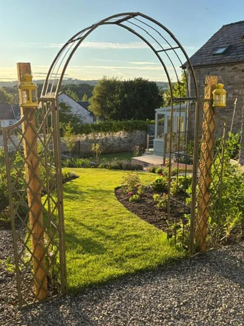 Metal garden archway with decorative lanterns, opening to a lush, well-maintained backyard with green grass and flower beds under a clear sky.