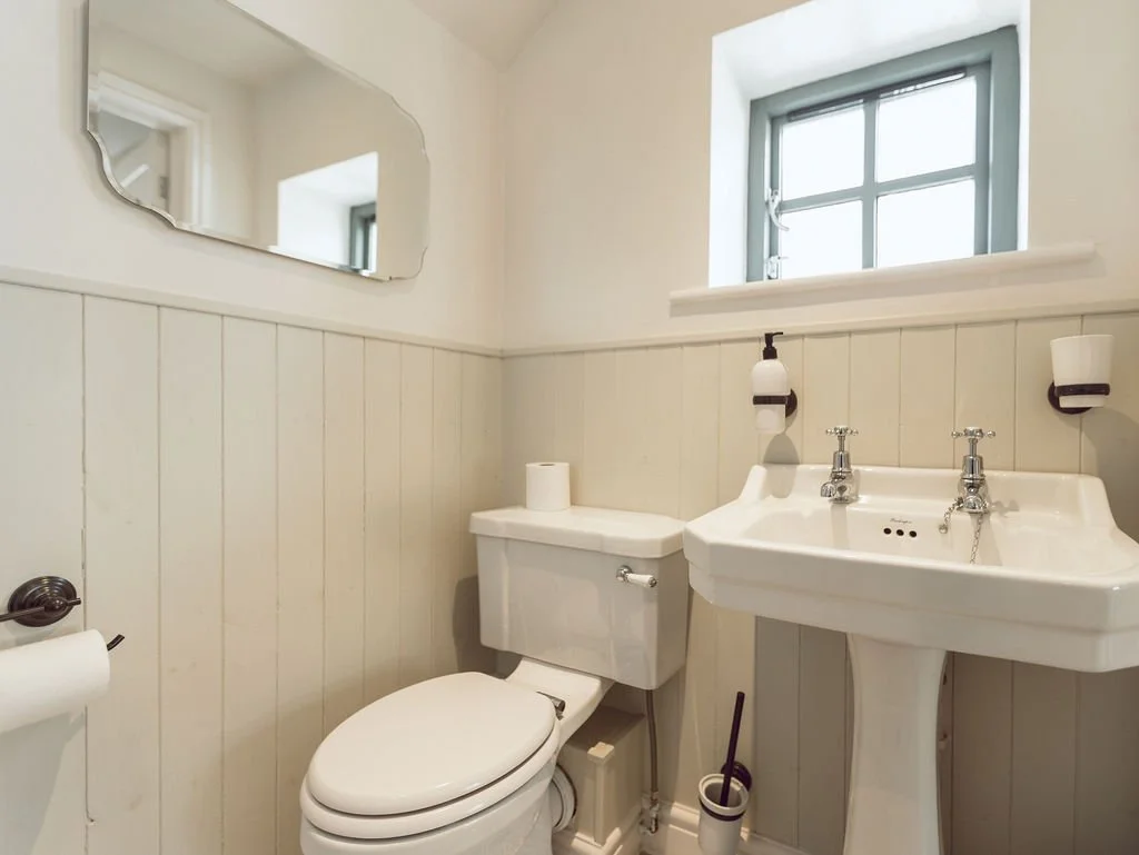 A small bathroom with a toilet, a pedestal sink, a mirror, a window, and wall-mounted light fixtures.