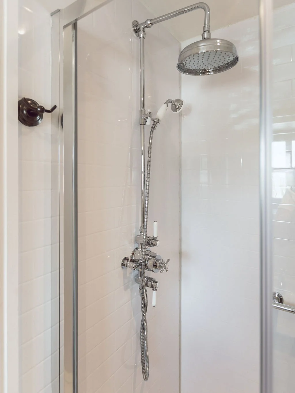 Shower with a chrome showerhead and handheld shower wand, enclosed by glass walls, with white tiled walls.