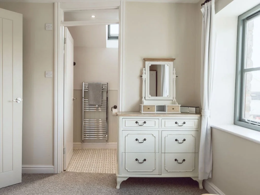 A white dresser with a mirror and small drawers in a bright room with a window and curtains, and an adjacent bathroom with a towel hanging on a heated towel rail.