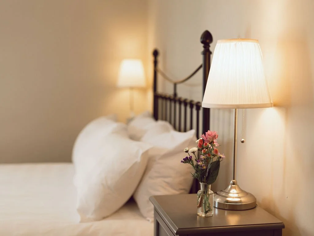 A bedroom with a bed, pillows, a nightstand, a lamp, and a small vase of flowers.