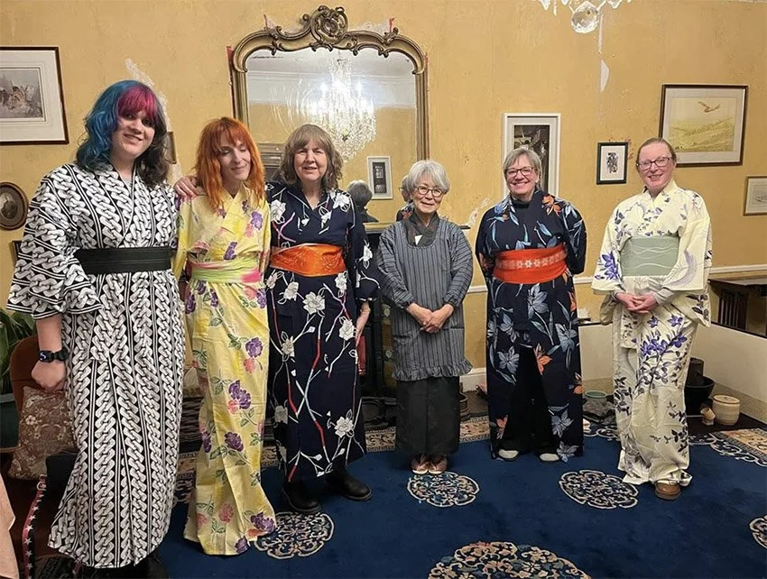 Six women standing together in a decorated room, some wearing traditional Japanese kimonos and one in a striped outfit, posing for a group photo.