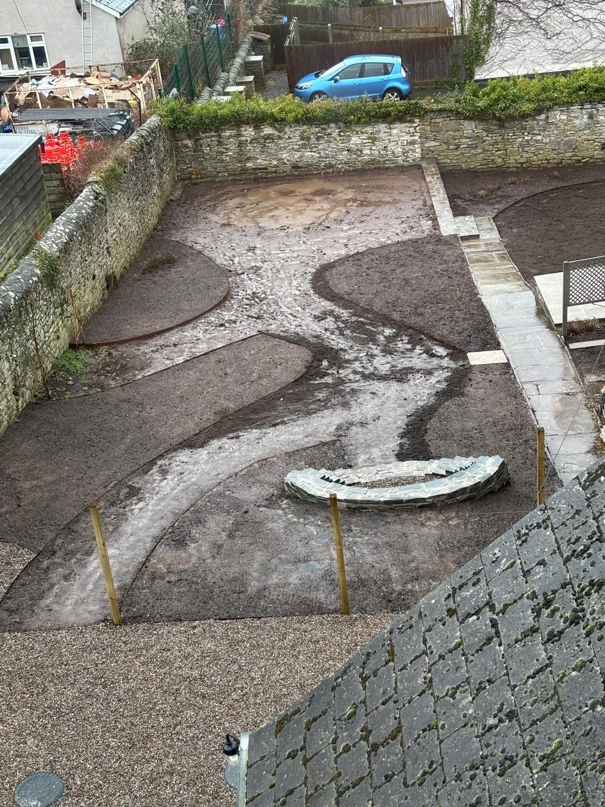 A backyard under construction with a winding pathway, some sections of the ground prepared for paving or landscaping, a stone wall at the back, and a blue car parked above the wall. Wooden stakes mark areas of the yard.