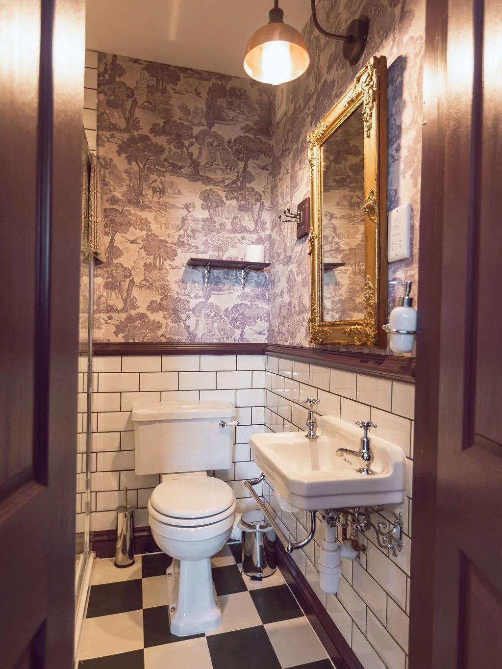 Vintage-style bathroom with black and white checkered floor, white toilet, small wall-mounted sink, ornate gold-framed mirror, patterned wallpaper, and warm lighting.