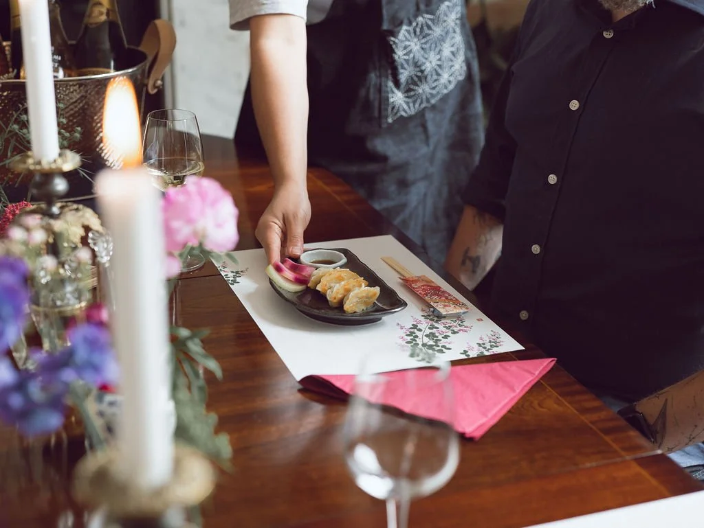 A person serving a plate of sushi with soy sauce on a black dish at a decorated dining table, with candles, flowers, wine glasses, and a paper napkin.