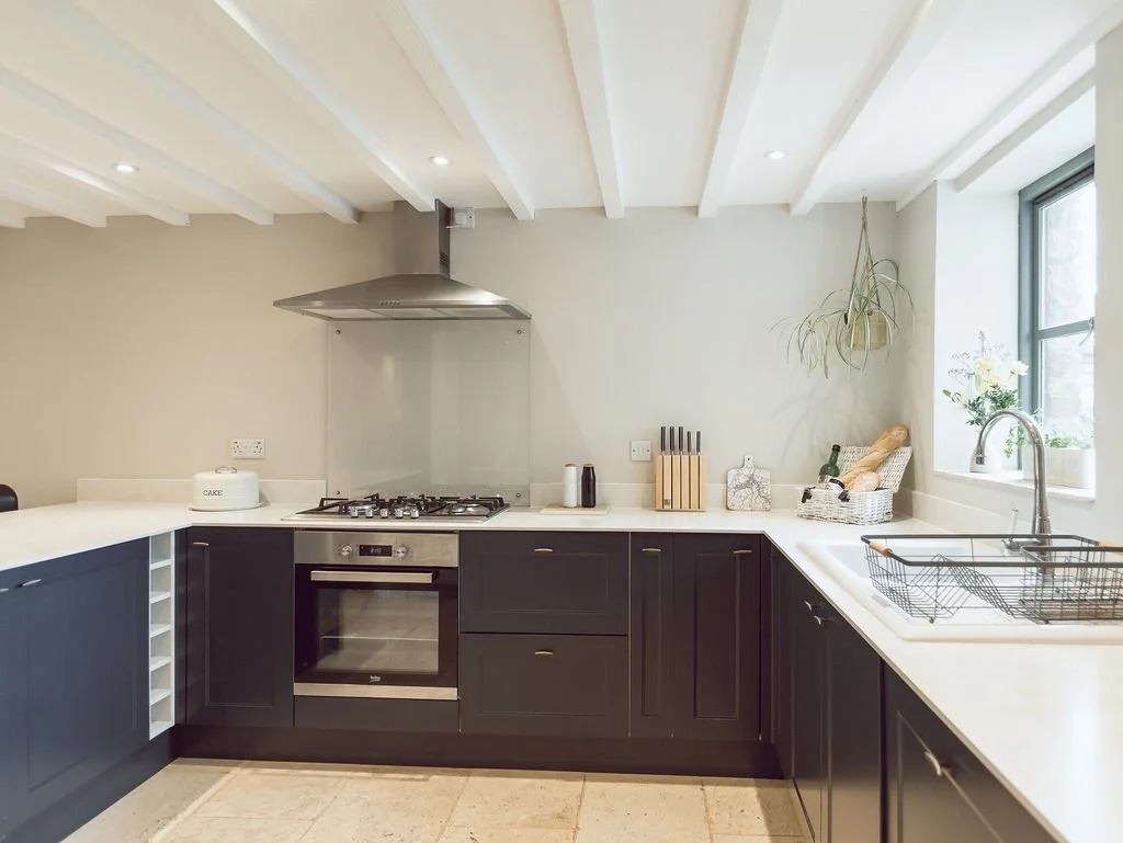 Modern kitchen with black lower cabinets, white countertops, stainless steel oven and range hood, a sink with dish rack, and a window with plants.