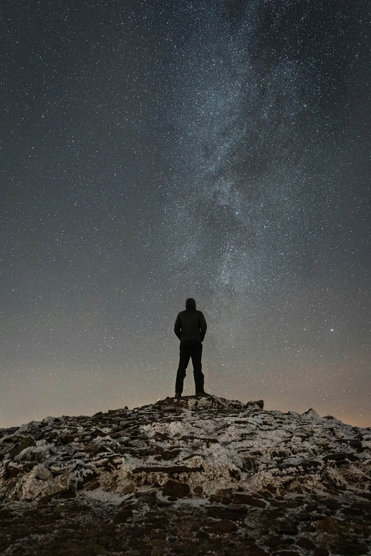 A person standing on a rocky, snow-covered hill, looking up at the starry night sky filled with countless stars and the Milky Way galaxy.