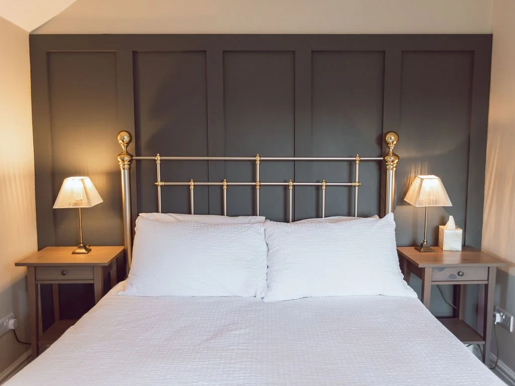 A neatly made bed with white bedding and two pillows, flanked by two wooden nightstands with lamps, in front of a dark-colored paneled wall with a brass bed frame.