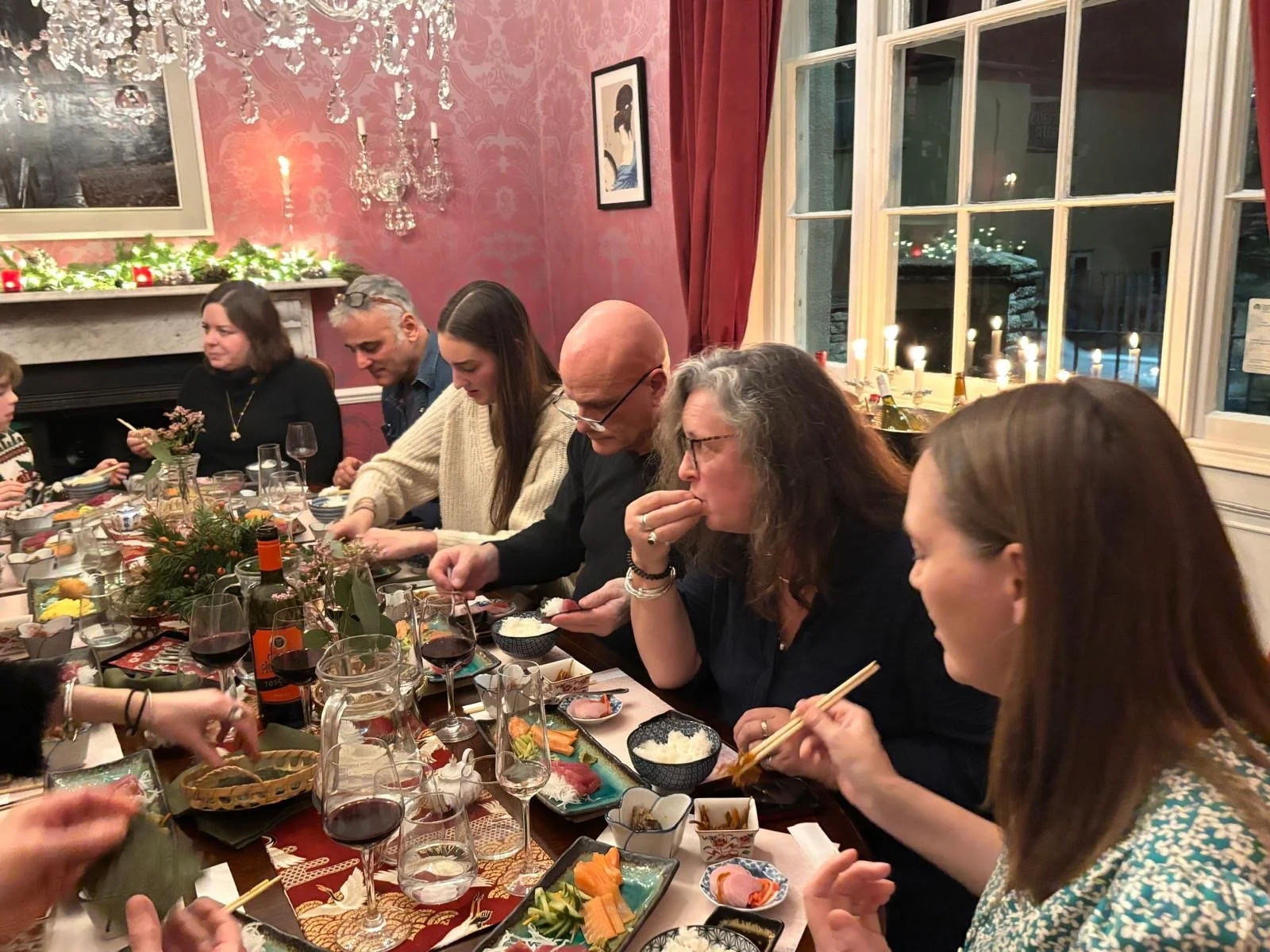 People sitting at a long dinner table enjoying a meal in a decorated dining room with pink wallpaper, candles, and a fireplace.