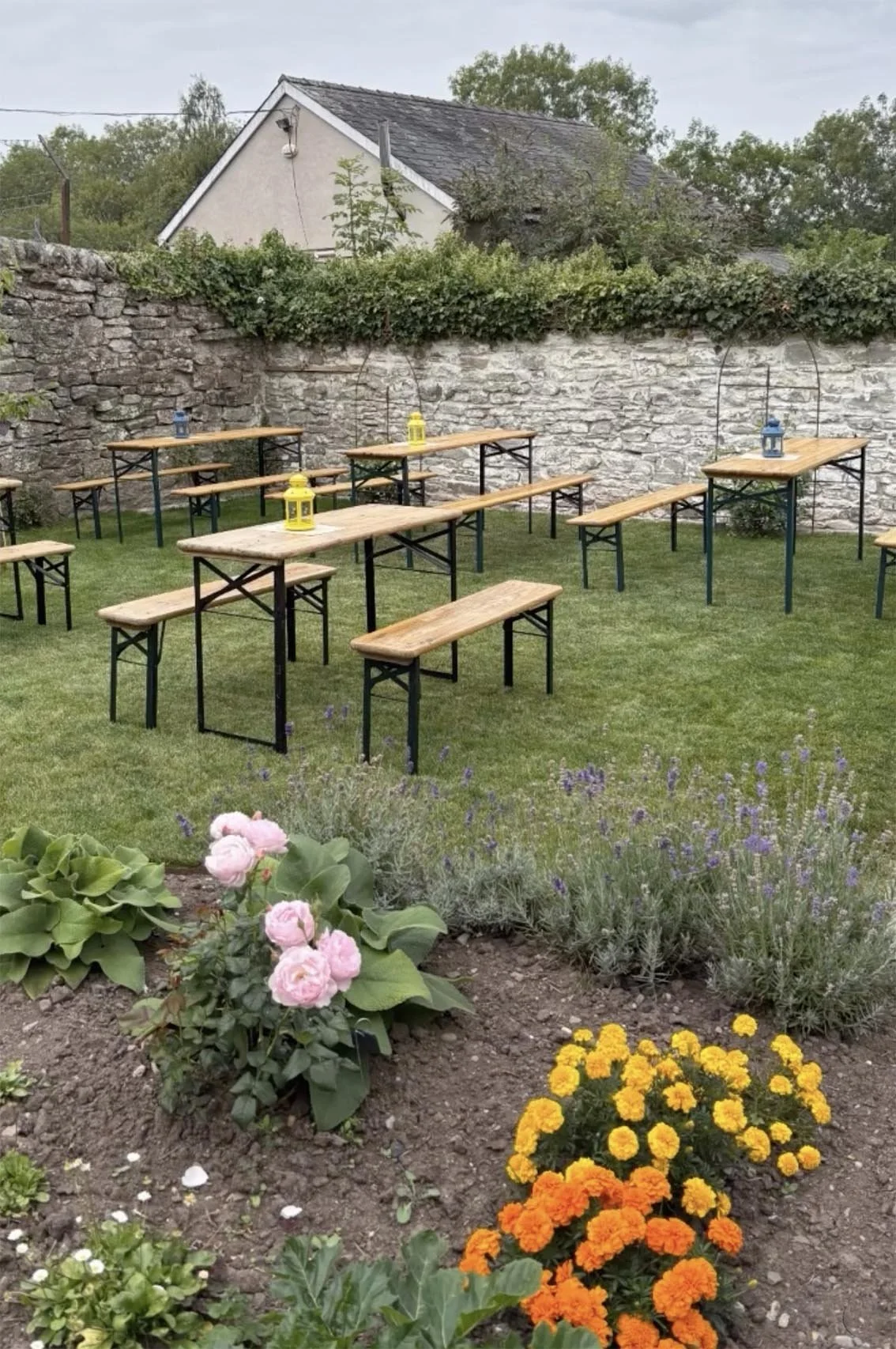 Outdoor garden with wooden tables and benches, decorated with lanterns, set against a stone wall, with flowers and plants in the foreground.