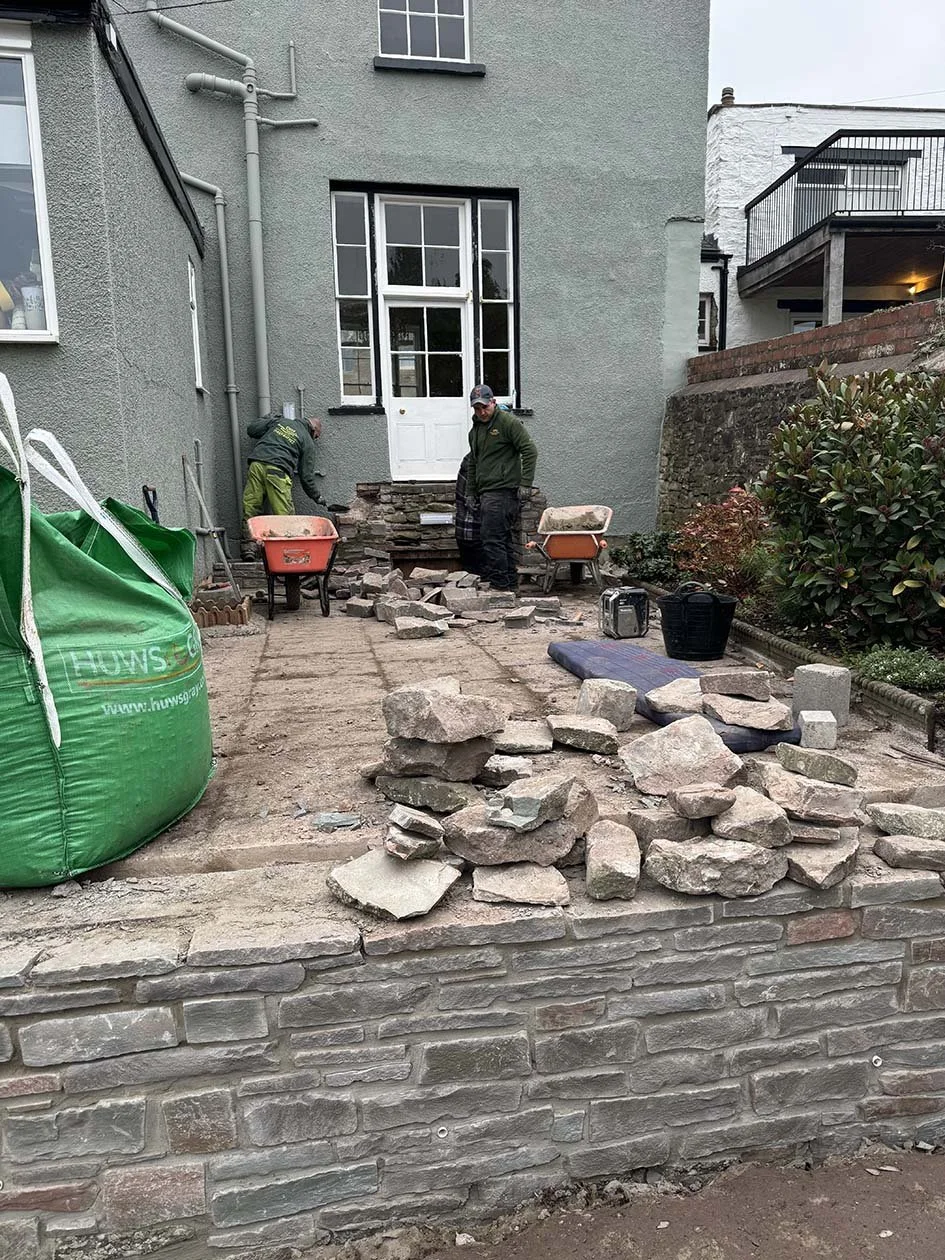 Two workers repairing a stone or brick wall outside a house with a large window and door. Construction materials and tools are visible around them.